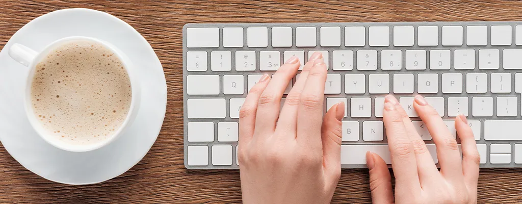 hands typing on a computer keyboard, with cup of coffee next to it